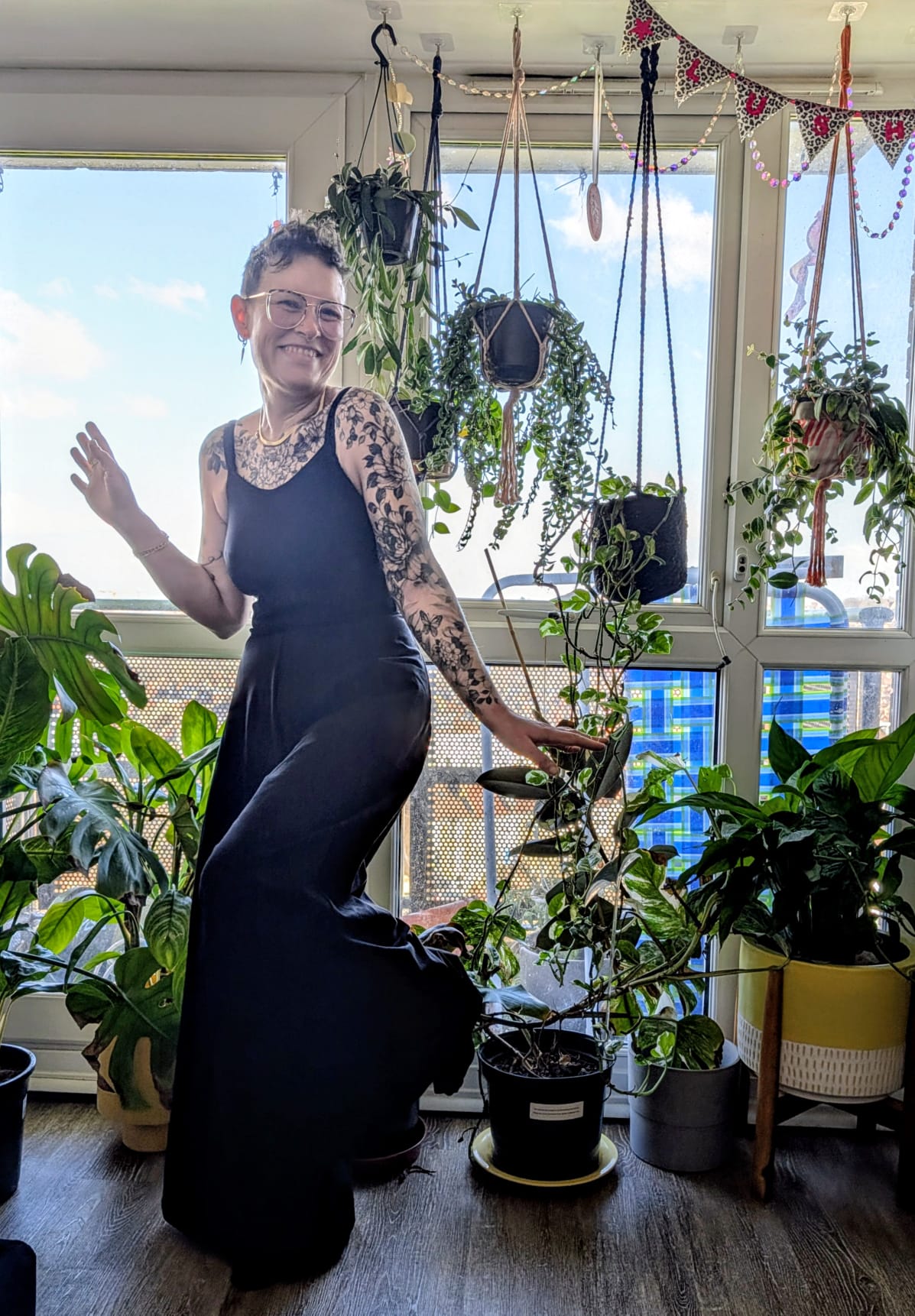 White non-binery person, stood in front of bright windows, surrounded by plants, in a playful pose